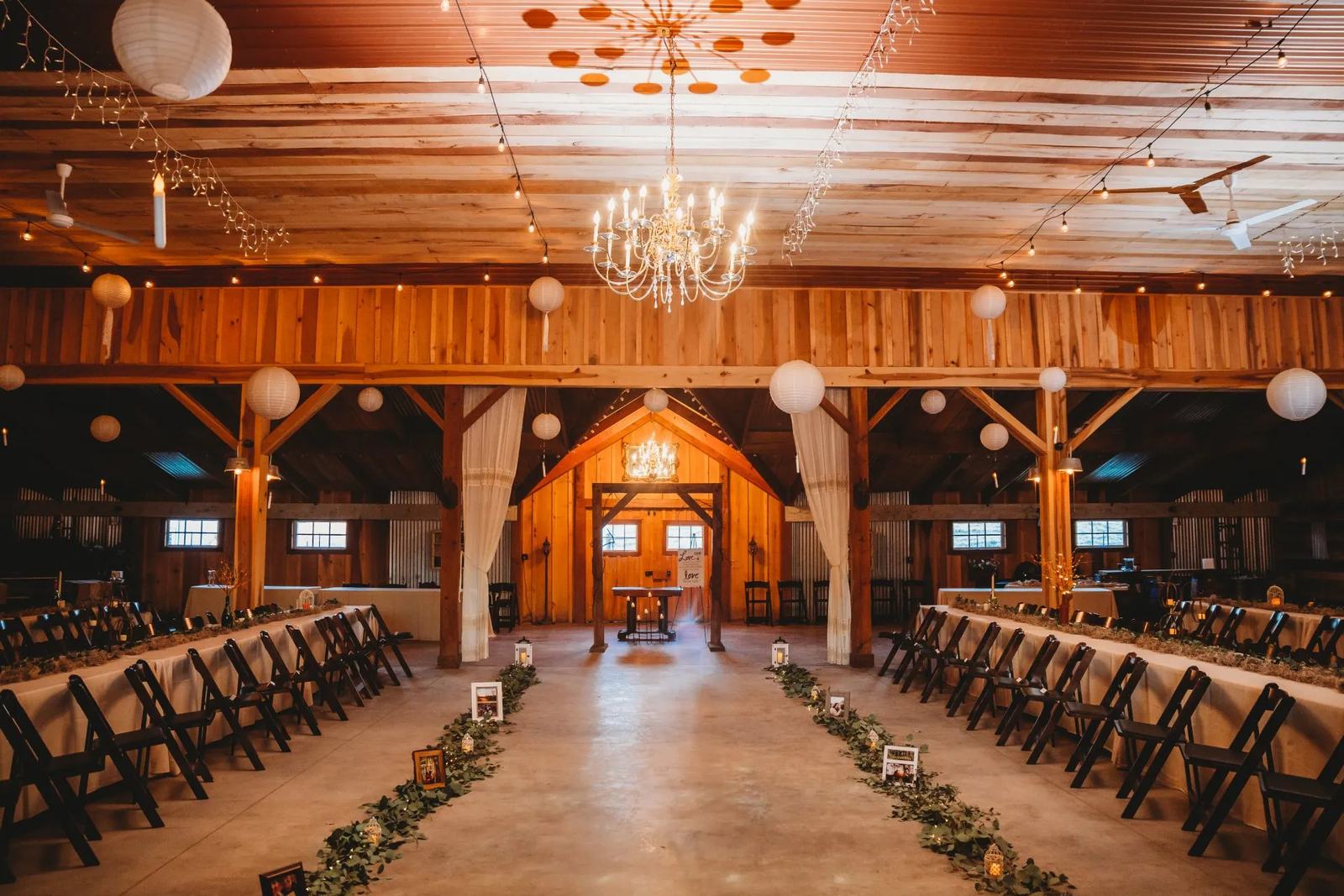 Inside the barn set for a wedding, with a chandelier, paper lanterns, string lights, long tables and a greenery-lined aisle