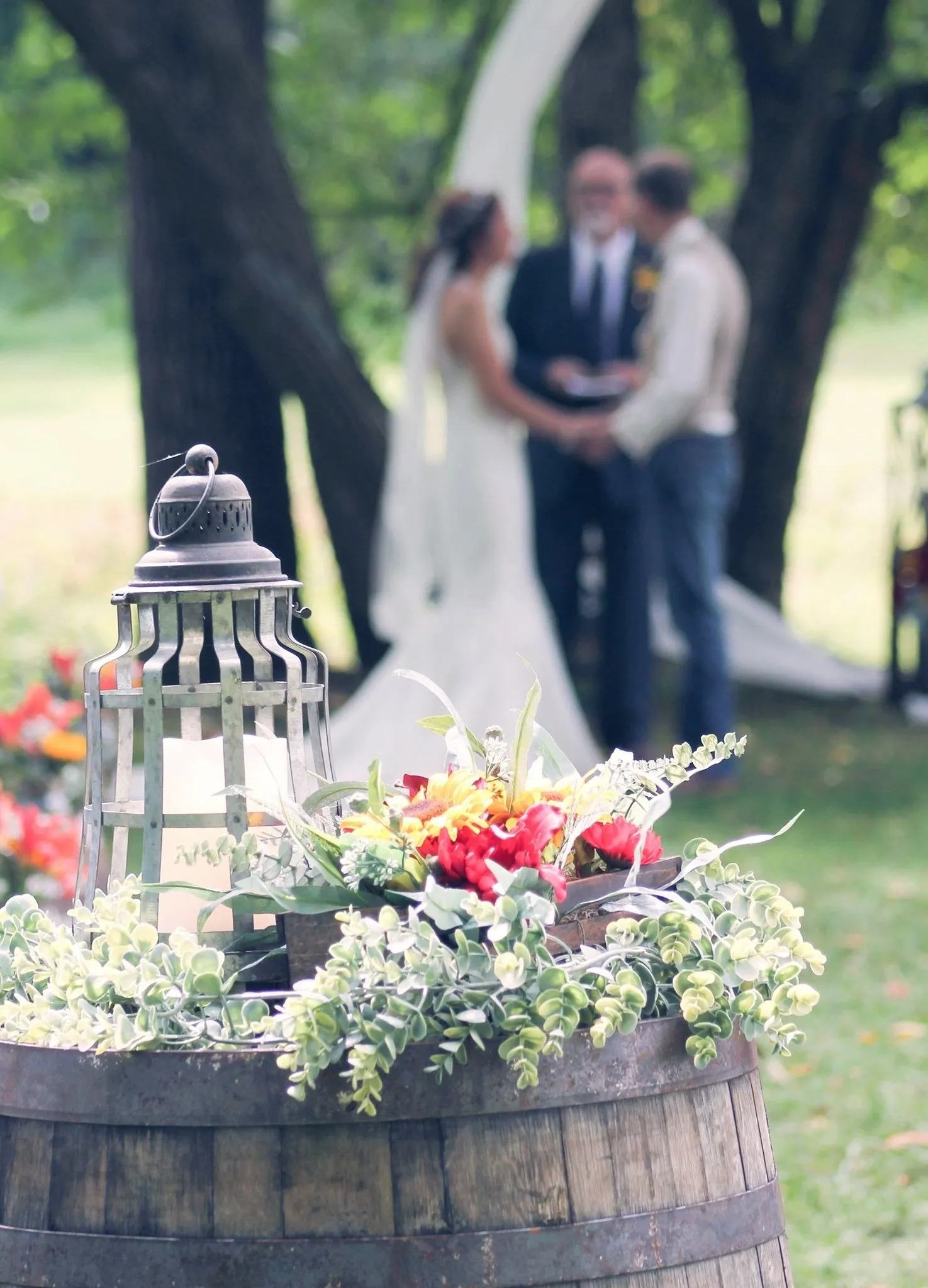Lantern and flowers on a whiskey barrel in the foreground, with a couple and officiant at the ceremony under the trees behind