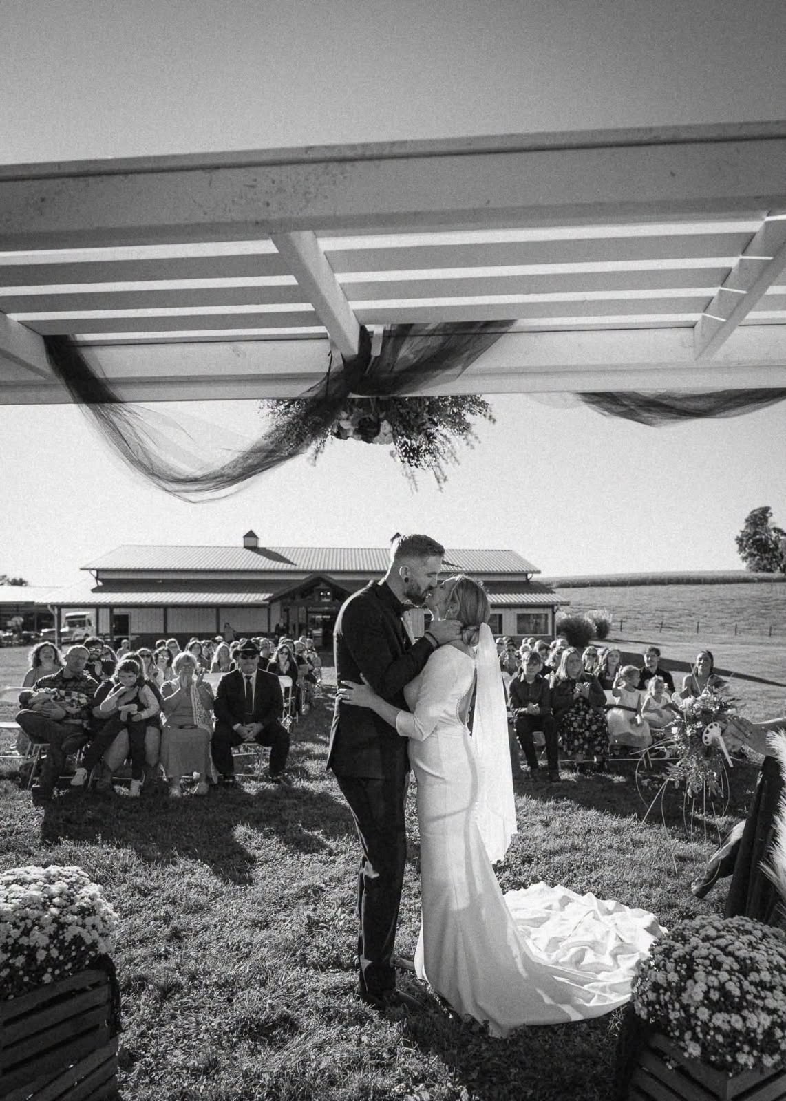 Black and white photo of a bride and groom kissing under the white pergola at their outdoor ceremony as guests applaud, with the event barn behind