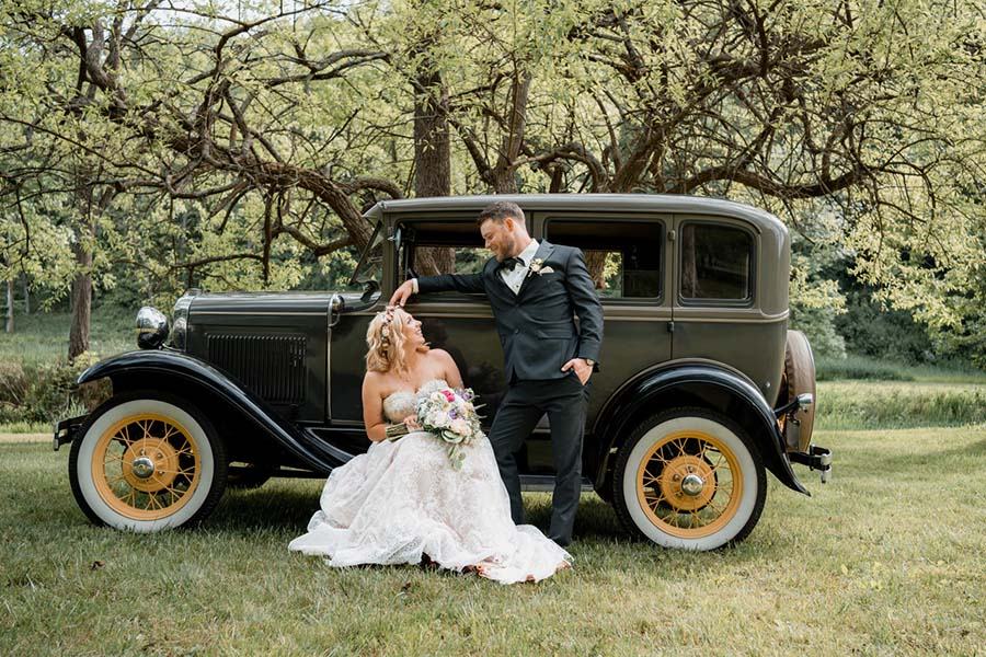 Bride sitting on the running board of a vintage car, smiling up at the groom