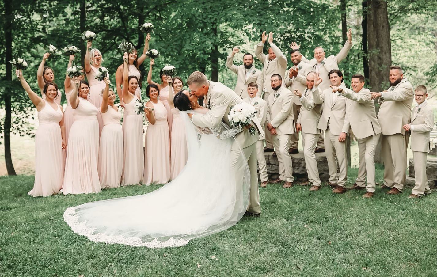 Groom dipping the bride for a kiss while the wedding party cheers under the trees